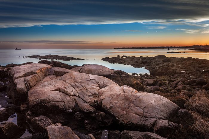 Boulders On The Shore At Sunset With Lighthouse In The Distance