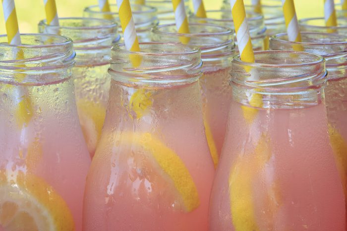 Close up of picnic party in the park drink table with large pitcher and tops of glass bottles filled with ice cold pink lemonade and fresh lemons with yellow swirled straws