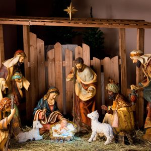 Figurines surround a baby in a manger, depicting a nativity scene, under a wooden shelter with straw and lambs, surrounded by trees.