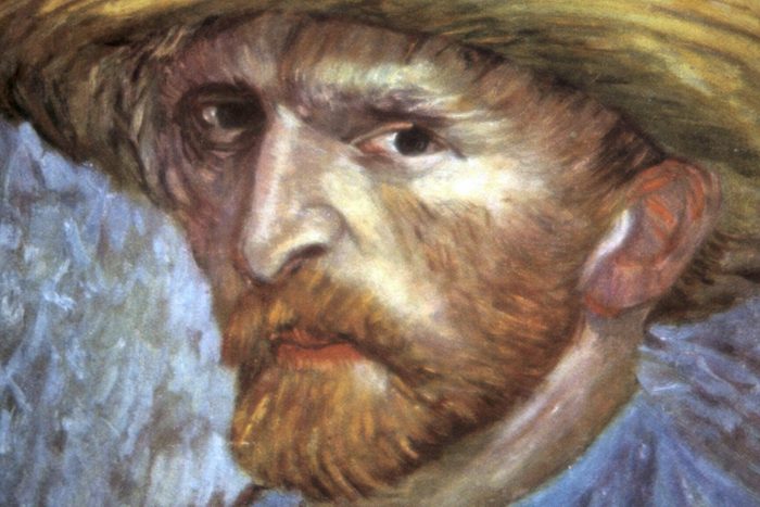 A man with a beard wears a yellow hat, gazing intensely. The background is textured with muted colors, enhancing the portrait’s focus.
