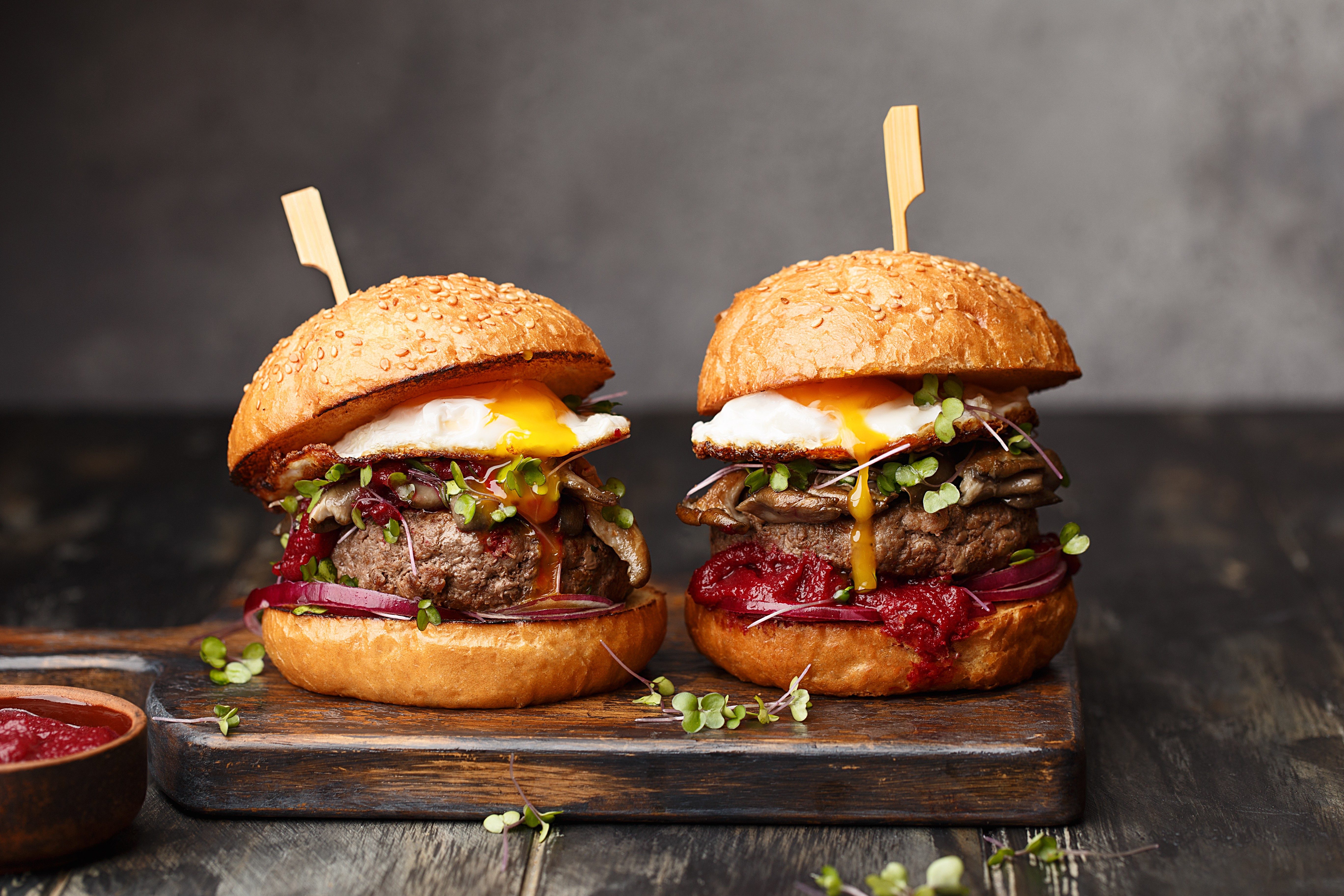 Two homemade beef burgers with mushrooms, micro greens, red onion, fried eggs and beet sauce on wooden cutting board