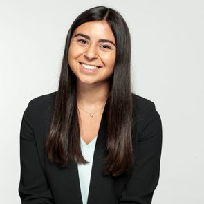 A person smiles seated, wearing a black blazer, in a brightly lit studio with a plain, light background.
