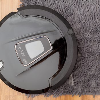 Robot vacuum cleaning, partially on a gray textured rug, with wood flooring visible.