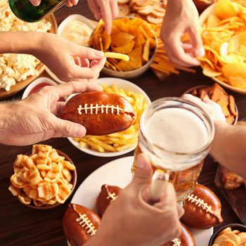 Hands share snacks, including football-shaped bread and chips, around a table with popcorn, fries, and drinks, creating a lively party atmosphere.