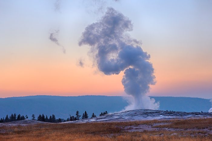 Steam rises from a geyser at sunset, surrounded by grassland and trees, with a hazy sky transitioning from orange to blue.