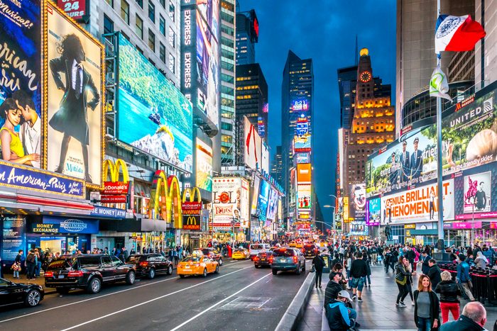 Billboards illuminate, cars drive, and people walk in a busy city square filled with lights and advertisements.