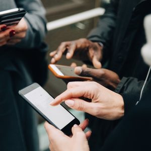 Three smartphones, held by hands, are used in close proximity, possibly for texting or browsing, in a social or public setting.