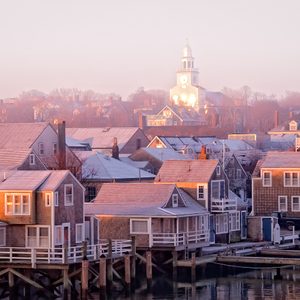 Snow-dusted rooftops reflecting sunrise glow, clustered around a waterfront community with a prominent church tower in the distance amidst a tranquil morning atmosphere.
