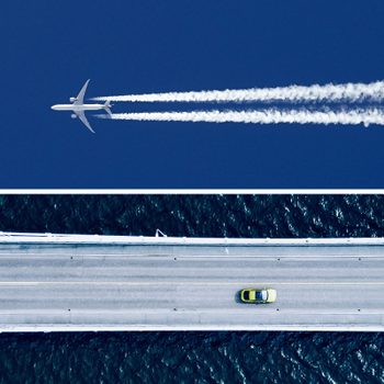 Airplane flies high releasing contrails in a clear blue sky; below, a yellow car drives on a bridge over dark water.