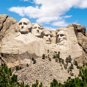 Rock sculptures depict four faces carved into a mountain, standing against a backdrop of blue sky and surrounded by evergreen trees.
