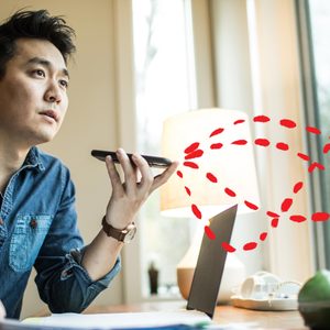 Man speaks into phone; digital signals depicted; seated at desk near a window with a laptop and lamp.