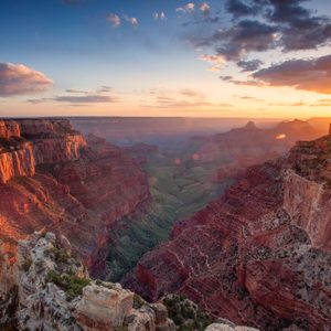 Canyon cliffs glow in the sunset light, surrounded by layered rock formations and a vast horizon under a partially cloudy sky.
