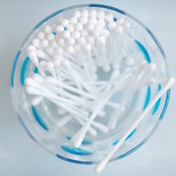 Cotton swabs rest in a clear glass jar, viewed from above, on a light surface, some swabs leaning and others upright.