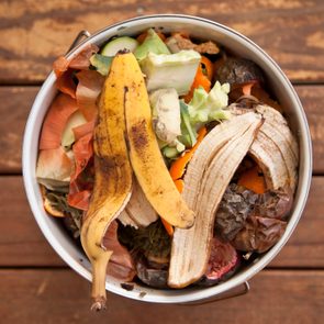 Compost bin holds assorted vegetable peels and banana skins, resting on a wooden deck.