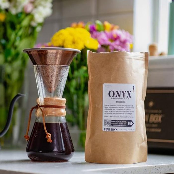 Coffee dripper brews on counter, next to Onyx Coffee Lab bag. Colorful flowers and kitchen items in the background.