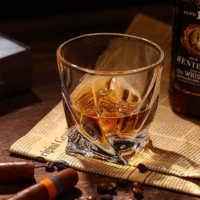 A glass holds amber liquid, possibly whiskey, on a newspaper with cigars and coffee beans, suggesting a relaxed, indulgent setting.