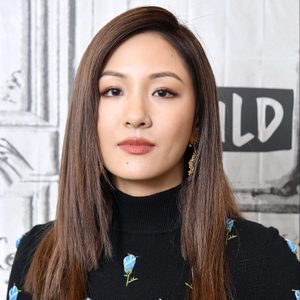 A woman with long, straight hair stands against a patterned gray wall, wearing a black turtleneck adorned with embroidered blue flowers.