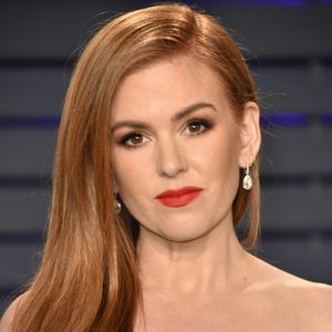 A woman with long red hair and red lipstick looks forward, wearing drop earrings, in front of a blurred dark striped background.