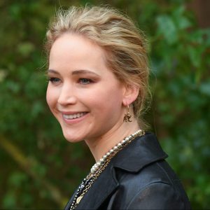 Person smiling outdoors, wearing a black leather jacket with layered necklaces and earrings; green, leafy background.