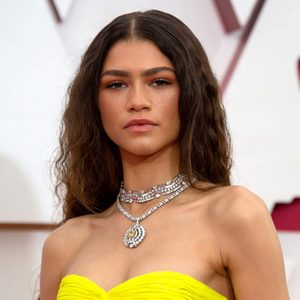 Woman wears bright yellow strapless dress and layered diamond necklace, standing confidently on a red carpet backdrop.
