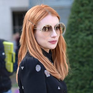 A woman with long red hair wears large round sunglasses, standing outdoors in a garden-like area.
