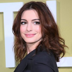A person with wavy brown hair smiles, wearing a black outfit, in front of a light-colored wall.
