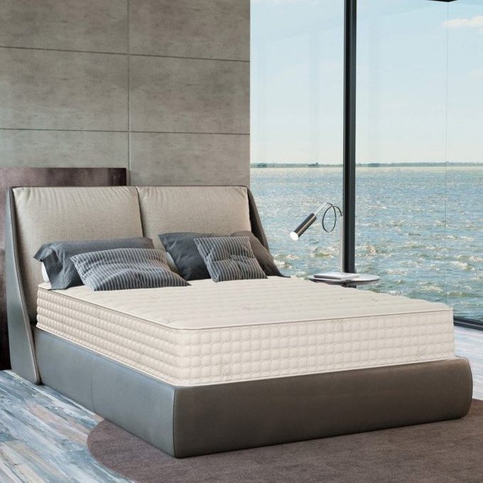 A modern bed with cushions sits in a luxurious bedroom by large windows overlooking a serene waterfront view.