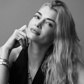 A woman with long, wavy hair poses thoughtfully, resting her chin on her hand, wearing a sleeveless top and jewelry, in a studio setting.