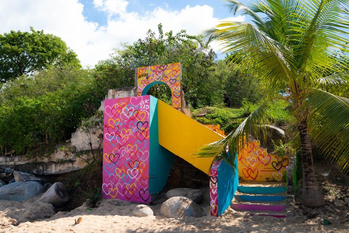 Colorful heart-covered structure stands on a sandy beach, surrounded by lush greenery and palm trees under a partly cloudy sky.