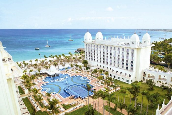 A large hotel overlooks an intricate pool area, framed by palm trees, beside a calm, turquoise ocean with several small boats visible.