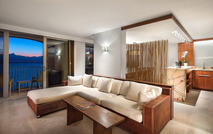 L-shaped couch sits adjacent to a wooden table, facing a large sliding window overlooking an ocean view, in a contemporary living room.