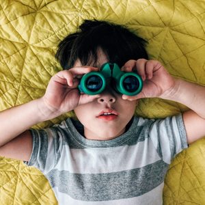 Child holds binoculars, lying on a yellow quilted blanket, wearing a gray and white striped shirt.