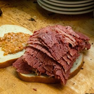 Sliced corned beef piled on rye bread with mustard, on a wooden cutting board, near a stack of white plates.