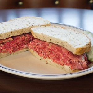 Sandwich filled generously with sliced deli meat, resting on a white plate, accompanied by a pickle slice in a dimly lit setting.