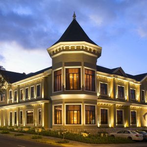A well-lit, elegant building with large windows and turret stands along a street, surrounded by greenery and parked cars, under a twilight sky.