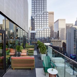 Rooftop terrace features colorful seating and planters, surrounded by glass railings, overlooking a cityscape with high-rise buildings.