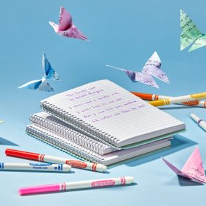 Colorful origami butterflies hover above stacked notebooks with handwritten text, surrounded by scattered Crayola markers on a light blue background.