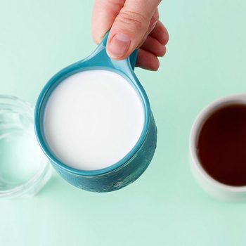 A hand pours milk from a blue cup into a glass, with a cup of coffee nearby on a light blue background.