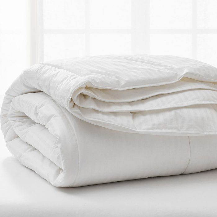 A neatly folded white comforter rests on a bed, in a bright room with diffused light from a window.