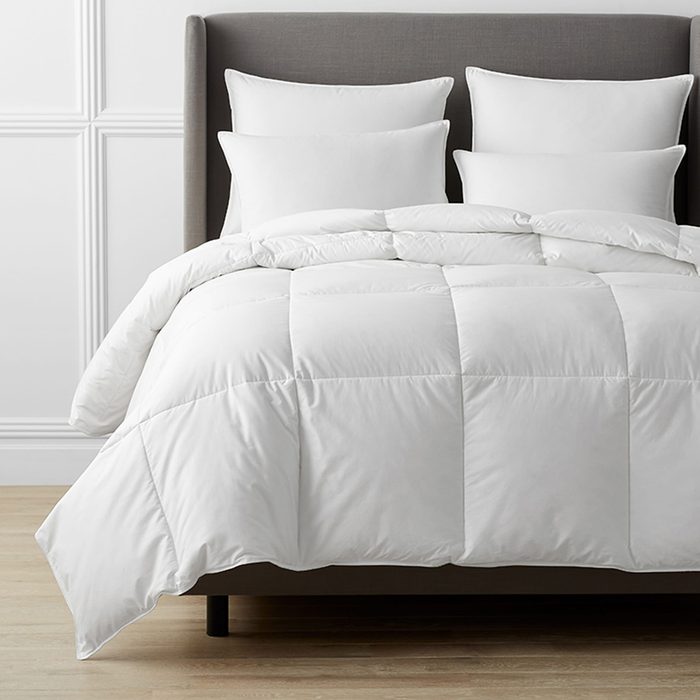 White comforter and pillows resting on a gray upholstered bed, placed in a minimalist bedroom with light-colored wooden flooring and white paneled walls.