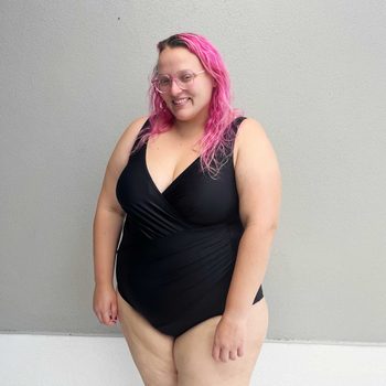 Person stands smiling, wearing a black swimsuit, against a plain light gray wall.