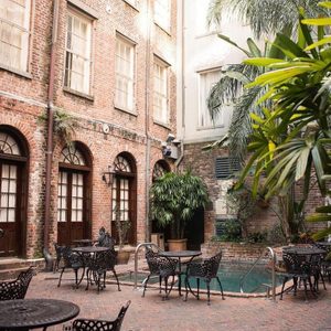 Wrought iron tables and chairs sit beside a small pool in a brick-walled courtyard with large plants and arched doors.