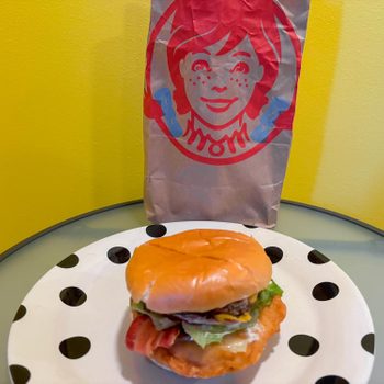Burger sits on polka-dot plate; behind, a brown paper bag with a red logo against a yellow wall.