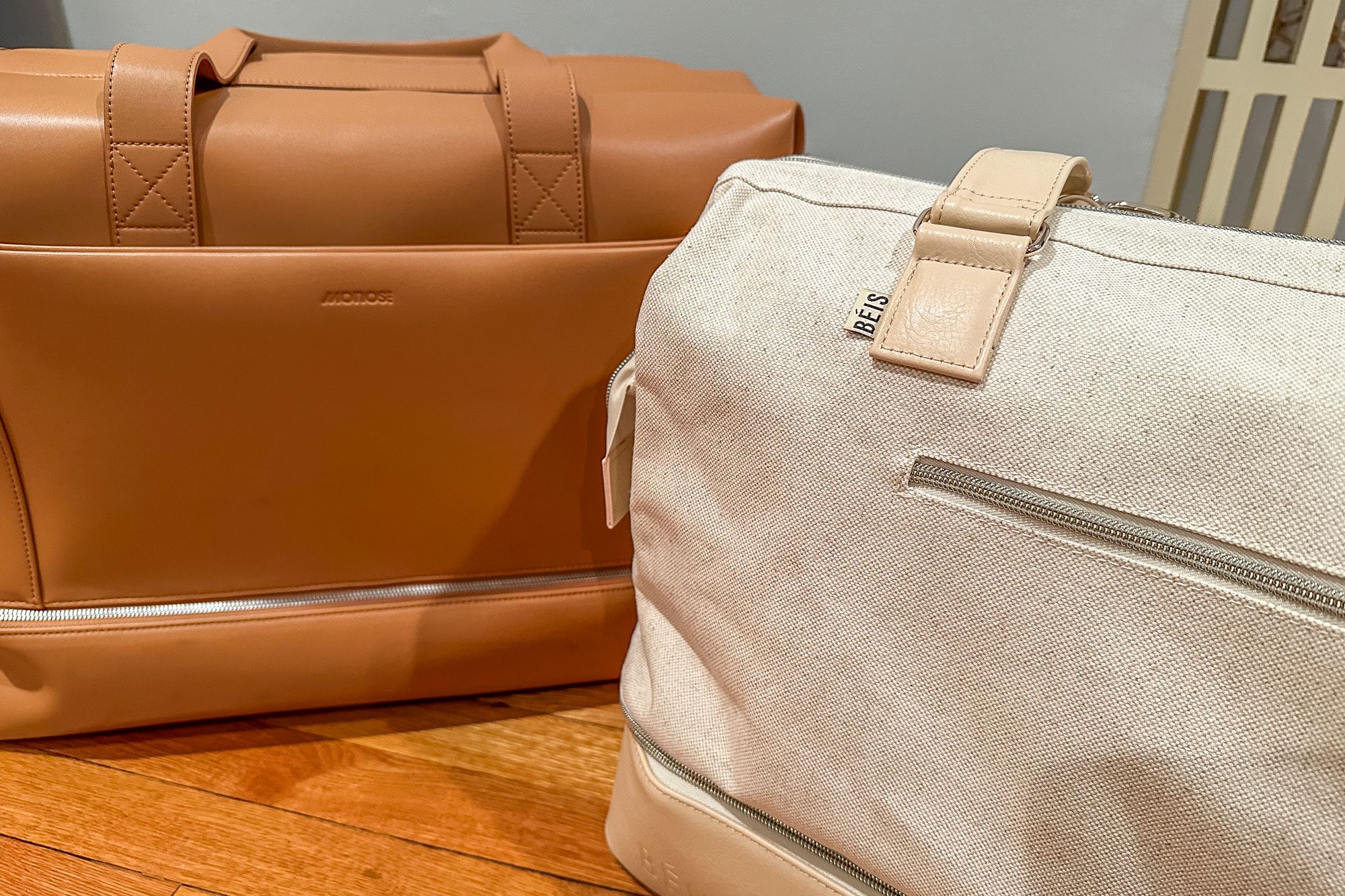 Two bags rest on hardwood floor; one is tan leather labeled "MONOS," and the other is cream fabric labeled "BÃ‰IS," featuring zippers.