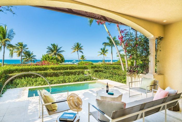 Patio overlooks pool and ocean, framed by palm trees. Chairs, table and hat suggest a relaxing tropical setting with clear skies.