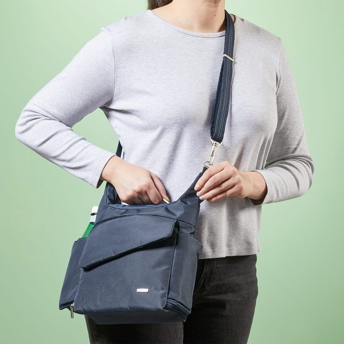 A person places a hand into a navy bag with pockets, against a green background, wearing a light grey shirt and dark pants.