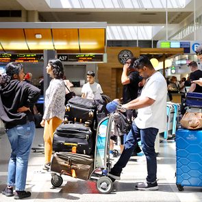 Travelers wait in line with luggage carts at an airport check-in area, surrounded by bright lighting and ticket counters displaying gates A37 to A40.