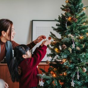 A woman and child decorate a lit Christmas tree with ornaments and pinecones in a cozy living room setting.