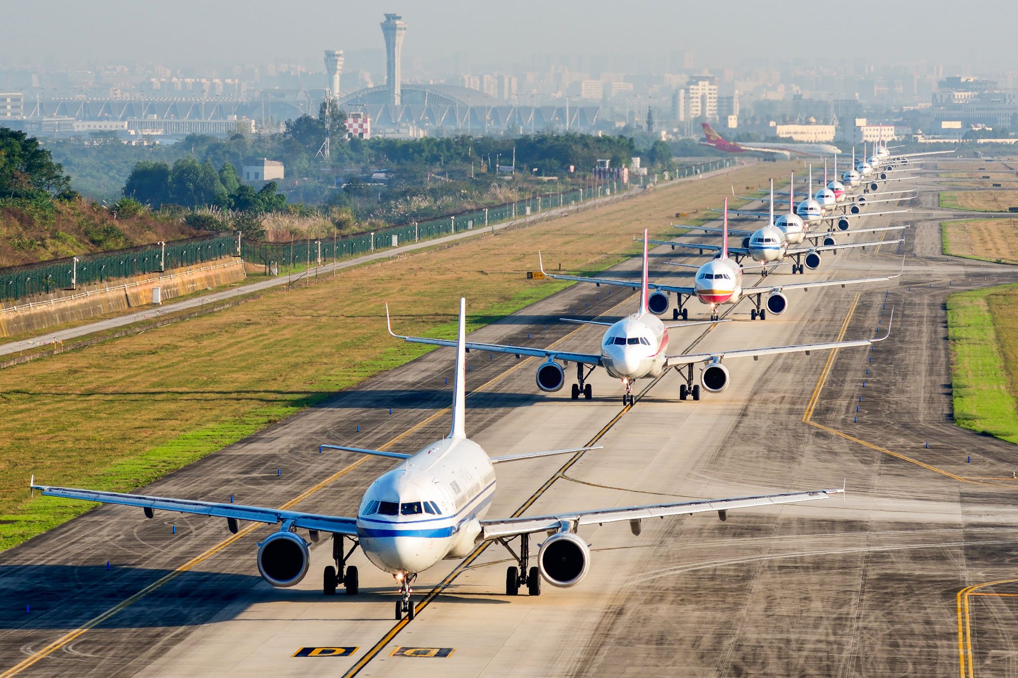 Airplanes lined up neatly on a runway, waiting to take off, surrounded by grassy fields and a distant cityscape.