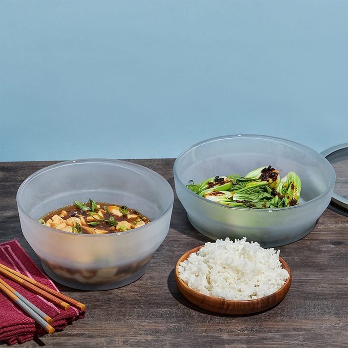 Two frosted bowls hold soup and vegetables; a wooden bowl with rice sits nearby on a wooden table, with chopsticks and napkins beside them.